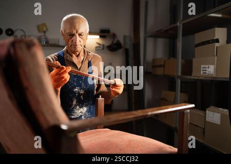 Älterer männlicher Möbelreparaturspezialist in Uniform entfernt hölzerne Armlehne von Vintage Stuhl restaurieren Sessel in der Werkstatt Stockfoto