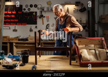 Älterer Restaurator entfernt die alte Schleifschicht des antiken Sessels mit einem speziellen Schleifpapierschwamm in der Werkstatt. Stockfoto