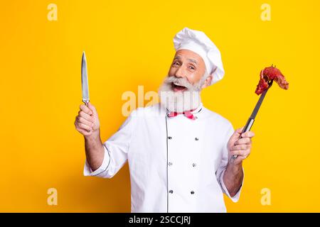 Chefkoch in Uniform, der Messer und Gabel mit Fleisch hält, vor einem leuchtend gelben Hintergrund Stockfoto