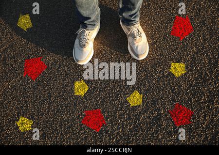 Mann, der neben roten und gelben Pfeilen steht, die in verschiedene Richtungen zeigen, auf Asphaltstraße, Nahaufnahme. Konzepte der Wahl, der Entscheidung, des alternativen Weges Stockfoto