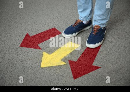 Mann, der neben roten und gelben Pfeilen steht, die in verschiedene Richtungen zeigen, auf Asphaltstraße, Nahaufnahme. Konzepte der Wahl, der Entscheidung, des alternativen Weges Stockfoto