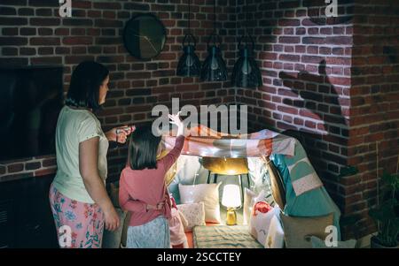 Rückansicht der Mutter, die eine Taschenlampe hält, während die Tochter Schattenpuppen mit Händen über der Wand macht. Stockfoto