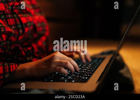 Nahaufnahme der Hände eines nicht erkennbaren Teenager-Mädchens mit rot-schwarzem kariertem Hemd, das auf der hinterleuchteten Laptop-Tastatur sitzt und in einem dunklen Zimmer auf dem Bett sitzt Stockfoto