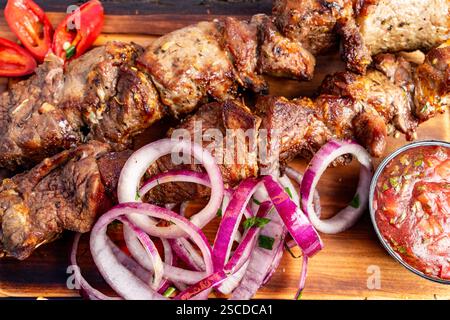 Saftige Grillspieße werden auf einer Holzplatte präsentiert, begleitet von frischen Zwiebeln und einer kleinen Schüssel scharfer Salsa. Farbenfrohe Chili-Paprika Stockfoto