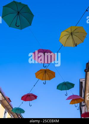 Farbenfrohe Regenschirme hängen in der Luft vor einem klaren blauen Himmel und schaffen eine lebendige und künstlerische Darstellung über einer Straße, die von Gebäuden gesäumt ist. Stockfoto