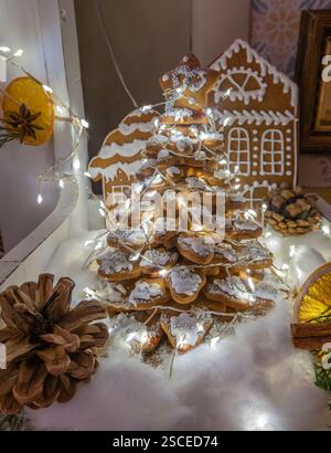 Ein festlicher Lebkuchenbaum, der mit Glasur verziert und von Lichtern umgeben ist, auf einer verschneiten Oberfläche mit einem Lebkuchenhaus im Hintergrund Stockfoto