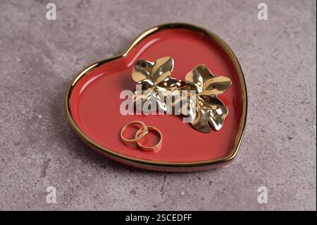 Ein paar goldene Hochzeitsringe und Ohrringe auf roter Herzform Keramikplatte mit goldenem Rand. Grauer Betonhintergrund. Valentinstag, Ehe Stockfoto