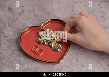 Ein paar goldene Hochzeitsringe und Ohrringe auf roter Herzform Keramikplatte mit goldenem Rand. Die Hand der Frau berührt Schmuck. Grauer Betonhintergrund Stockfoto
