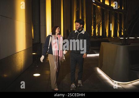 Ein lächelndes Paar, das nachts Hand in Hand in Hand geht in einer Stadt mit hellen Lichtern Stockfoto