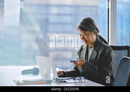Geschäftige asiatische Geschäftsfrau mittleren Alters, die ihr Mobiltelefon benutzt und im Büro arbeitet. Stockfoto