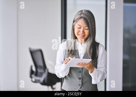 Lächelnde, reife asiatische Geschäftsfrau, die im Büro ein Tablet benutzt. Stockfoto