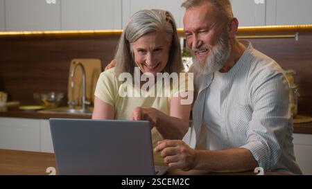 Familiengespräch mittleren Alters Verwenden Sie Laptop in der Küche, durchsuchen Sie soziale Netzwerke, buchen Sie Tickets Online-Einkäufe Lebensmittel Lieferung lächelnde Kaukasier Mann und Frau kaufen Stockfoto