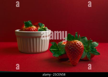 Eine Schüssel Erdbeeren ist auf rotem Hintergrund. Auf dem Tisch liegen drei Erdbeeren. Die Schüssel ist weiß Stockfoto