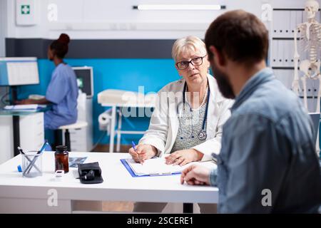 Pensionierte Ärztin mit Klemmbrett-Beratung mit jungen männlichen Patienten in der Klinik als Krankenschwester im Hintergrund. Älterer Arzt hört Mann mit Bart zu und schreibt seine medizinischen Symptome. Stockfoto