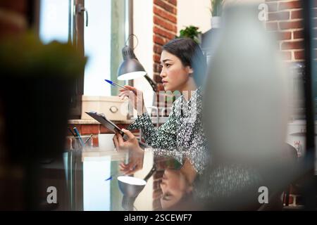 Eine Forscherin analysiert Geschäftsanalysen auf ihrem Computerbildschirm und konzentriert sich dabei auf die Unternehmensdaten. Asiatische Geschäftsfrau studiert Finanzdiagramme auf Desktop-pcs und hält einen Stift und eine Zwischenablage. Stockfoto
