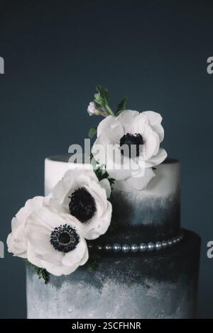 Ein wunderschön gefertigter Kuchen steht hoch und hat glatte Schichten in dunklen Tönen. Weiße Blüten mit schwarzen Mittelpunkten kaskadieren an der Seite und verstärken ihre e Stockfoto