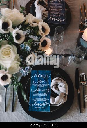 Der wunderschön gestaltete Hochzeitstisch verfügt über ein elegantes, florales Herzstück aus weißen Rosen und Anemonen sowie ein detailliertes Menü, das eine Einladung schafft Stockfoto