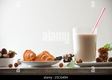 Glas Haselnussgetränk mit Strohhalm auf hölzerner Küchenbank mit Früchten und Schokolade und Croissants mit isoliertem Hintergrund. Vorderansicht. Stockfoto