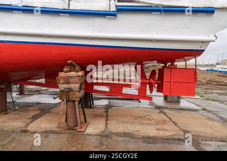 Trocken angedocktes Boot mit rot-weißem Rumpf, das Rudersystem und Propeller zeigt, gestützt von Holz- und Metallblöcken, in unmittelbarer Nähe der Werft Stockfoto
