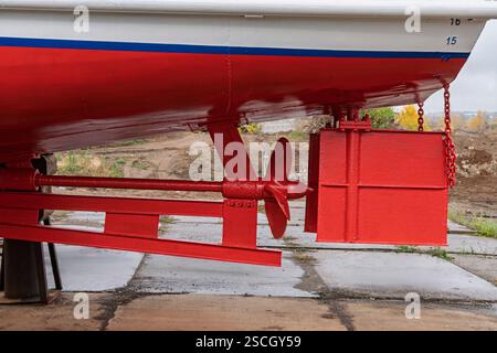 Trocken angedocktes Boot mit rot-weißem Rumpf, das Rudersystem und Propeller zeigt, gestützt von Holz- und Metallblöcken, in unmittelbarer Nähe der Werft Stockfoto