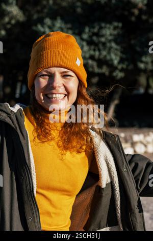 Fröhliche junge Frau in orangefarbener Mütze und Pullover, die draußen lacht Stockfoto