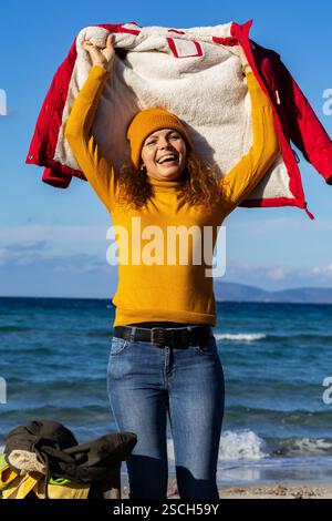 Fröhliche Frau, die am sonnigen Frühlingstag am Strand ihre Jacke hochhebt Stockfoto