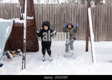 Zwei Kinder schwingen im Schnee auf einem Spielset im Garten mit fallendem Schnee Stockfoto