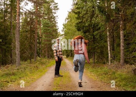 Ein Paar genießt einen gemütlichen Spaziergang auf einem unbefestigten Pfad in einem schwedischen Wald. Der Mann lächelt, während die Frau fröhlich überspringt, umgeben von üppigem Grün und Stockfoto