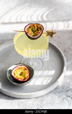 Eleganter Gin-Tonic-Cocktail, gemischt mit Passionsfruchtsaft, garniert mit einer frischen Passionsfrucht-Hälfte, präsentiert auf einem stilvollen grauen Tablett unter weicher Oberfläche Stockfoto