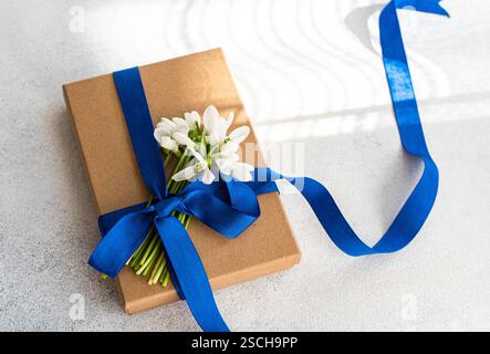 Blick von oben auf eine braune Geschenkbox, die mit einem lebhaften blauen Band und frischen Schneeglöckchen verziert ist, anmutig auf einem strukturierten weißen Hintergrund angeordnet. Stockfoto