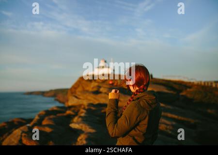 Rückansicht einer nicht erkennbaren Frau mit rotem Haar, die am Cape Spear Lighthouse in Kanada steht. Sie trägt eine grüne Jacke und blickt auf die Long Stockfoto