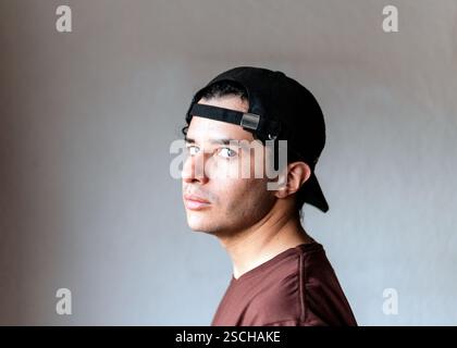 Ein junger Mann mit neutralem Ausdruck, der ein Headset und eine schwarze Kappe trägt, posiert vor grauem Hintergrund Stockfoto