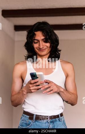 Ein junger Mann in einem weißen Tanktop und einer Jeans nutzt sein Smartphone während einer Pause beim Umbau zu Hause und zeigt ein lässiges, aber fokussiertes Verhalten. Stockfoto