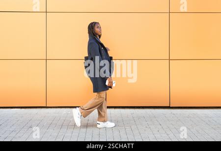 Eine junge afroamerikanische Geschäftsfrau mit Zöpfen, gekleidet in einer formellen grauen Jacke und beigefarbenen Hose, die ein Smartphone hält, während sie gegen ein V geht Stockfoto