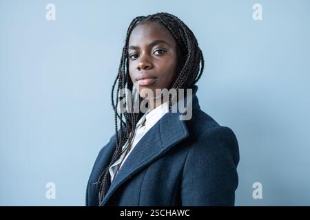 Afroamerikanische Geschäftsfrau mit Zöpfen, in einem formellen Anzug gekleidet, posiert vor blauem Hintergrund und spiegelt Professionalität und Selbstvertrauen wider. Stockfoto
