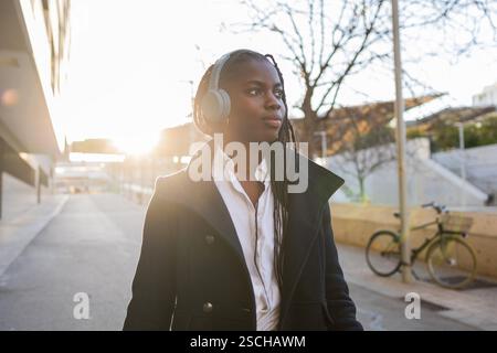 Eine afroamerikanische Geschäftsfrau mit Zöpfen macht draußen eine Pause, trägt Kopfhörer und einen stylischen Blazer über einem weißen Hemd. Stockfoto