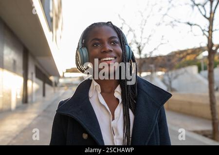 Eine lächelnde afroamerikanische Geschäftsfrau mit Zöpfen macht eine Pause im Freien und hört Musik über Kopfhörer. Stockfoto