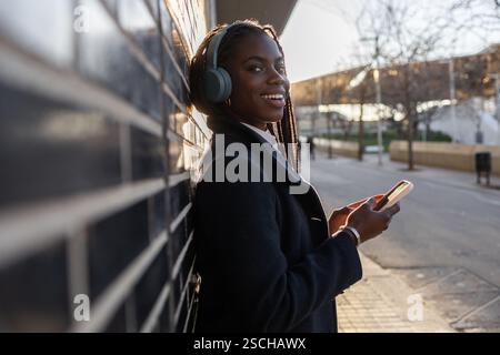 Eine afroamerikanische Geschäftsfrau mit Zöpfen lächelt, während sie im Freien eine Pause macht, Musik über Kopfhörer hört und ihr Smartphone benutzt. Stockfoto