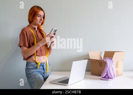 Eine Frau organisiert ihre Kleidung, um sie online zu verkaufen. Sie fotografiert mit ihrem Telefon Gegenstände und mit ihrem Laptop Angebote, um einen modernen Dekl zu illustrieren Stockfoto