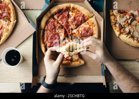 Von oben beschnittene, nicht erkennbare Hände, die ein Stück Pizza hielten, mit mehreren offenen Pizzaschachteln gefüllt mit verschiedenen Belägen. Die se Stockfoto