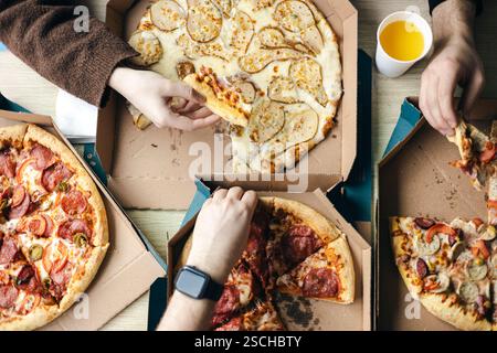 Blick von oben auf eine Gruppe nicht erkennbarer Freunde, die verschiedene Pizzen zusammen genießen. Verschiedene Hände, sowohl männlich als auch weiblich, greifen in Kartonschachteln zu gr Stockfoto
