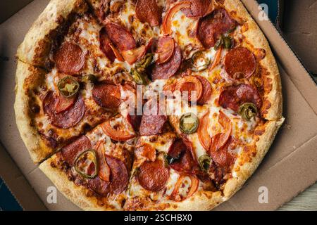 Blick von oben auf eine frisch gebackene Peperoni-Pizza mit Jalapenos, Tomaten und geschmolzenem Käse, serviert in einer Pappverpackung, betont die Temperatur Stockfoto