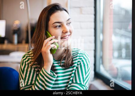 Eine fröhliche Frau genießt eine Kaffeepause in einem Café, während sie mit ihrem Smartphone plaudert, gekleidet in einem lässigen gestreiften Oberteil. Stockfoto