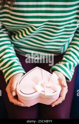 Abgeschnittene, nicht erkennbare Frau in einem Café, die eine herzförmige Geschenkbox hält, die mit einem weißen Band verziert ist. Das Bild konzentriert sich auf das Geschenk, das sie in den Händen hält Stockfoto