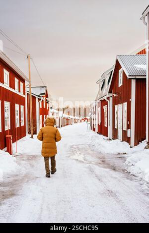 Ein einsamer Reisender in einem warmen Mantel spaziert durch ein bezauberndes verschneite Dorf in Schwedisch Lappland, umgeben von traditionellen roten Holzhäusern und einem ruhigen Sieg Stockfoto