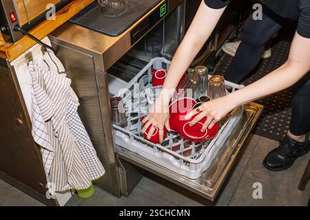Eine Person lädt einen Geschirrspüler mit roten Kaffeetassen und Glasflaschen, um die Vorgänge hinter den Kulissen in einem speziellen Café oder Café envi zu erfassen Stockfoto