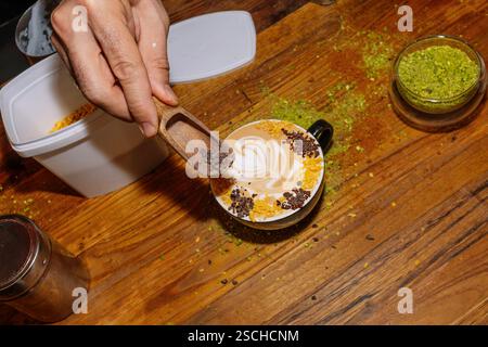 Eine Nahaufnahme einer Hand, die einen wunderschön gefertigten Kaffeespezialitäten mit komplizierter Latte Art ergänzt. Der Holztisch und das grüne Puder sorgen für ein ru Stockfoto