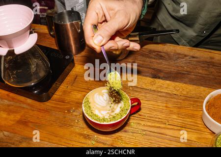 Ein Barista fügt Pistazienstaub zu einem wunderschön gefertigten Latte hinzu und zeigt die Kunst des Kaffeespezialitäten. Die Holztheke und die Kaffeewerkzeuge runden den ab Stockfoto