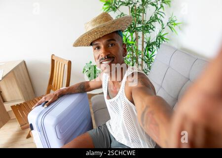 Eine fröhliche LGBTQIA+ Person mit Koffer, bereit für Urlaubsabenteuer. Sie tragen einen Strohhut und ein Mesh-Oberteil und versprühen Spannung und Urlaubs-spir Stockfoto
