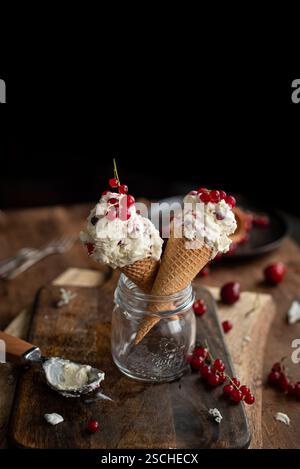Zwei Eiskegel gefüllt mit cremigem Dessert, garniert mit frischen Johannisbeeren, künstlerisch in einem Glasgefäß auf einem rustikalen Holztisch platziert, Begleiter Stockfoto
